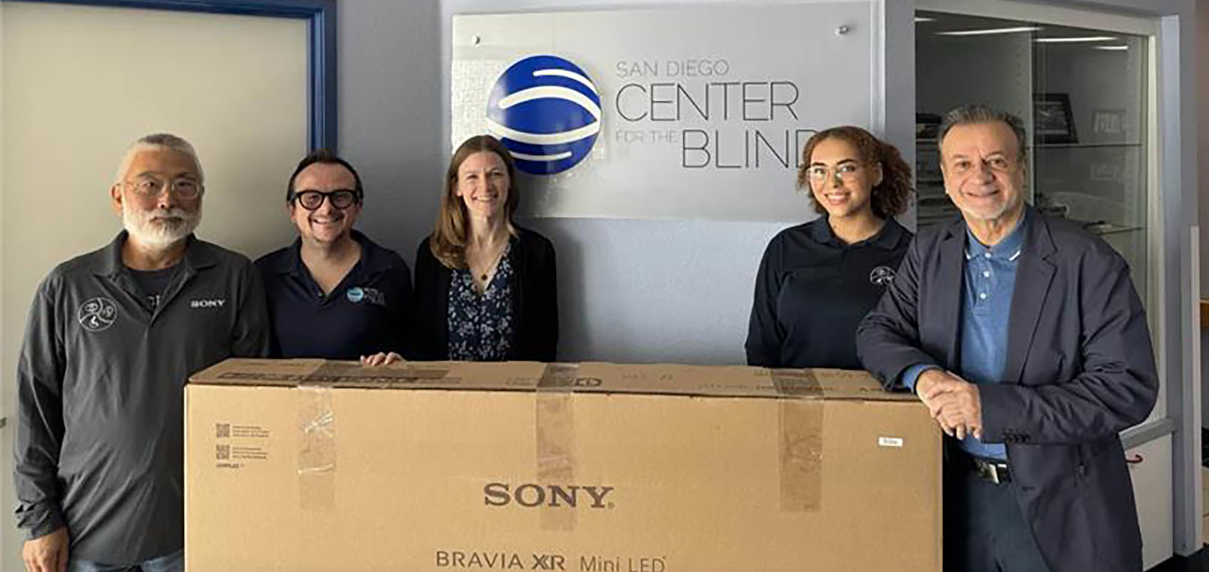 A group of people gathered around a BRAVIA box in front of a sign for the San Diego Center for the Blind.